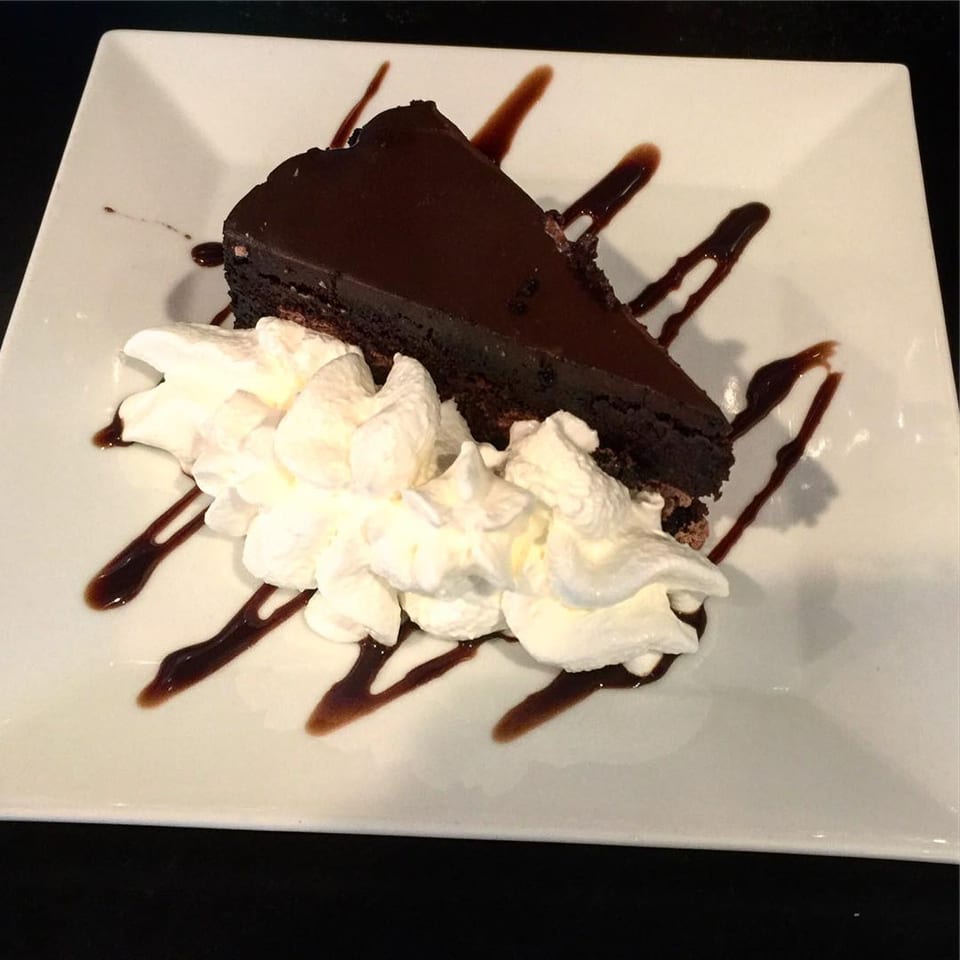 A slice of rich chocolate cake with whipped cream and chocolate drizzle on a white plate.