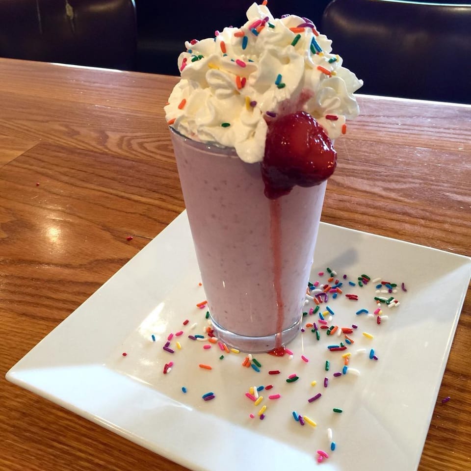 Strawberry milkshake with whipped cream, sprinkles, and a whole strawberry, on a white plate.