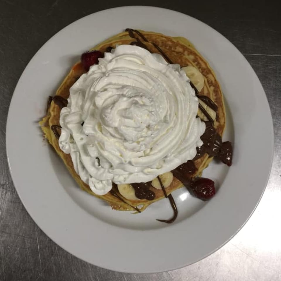 Diner pancakes topped with whipped cream, chocolate syrup, banana slices, and strawberries on a white plate.