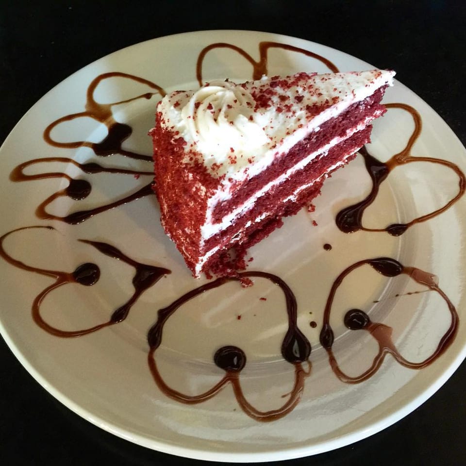 A slice of red velvet cake with white frosting and chocolate drizzle on a white plate.