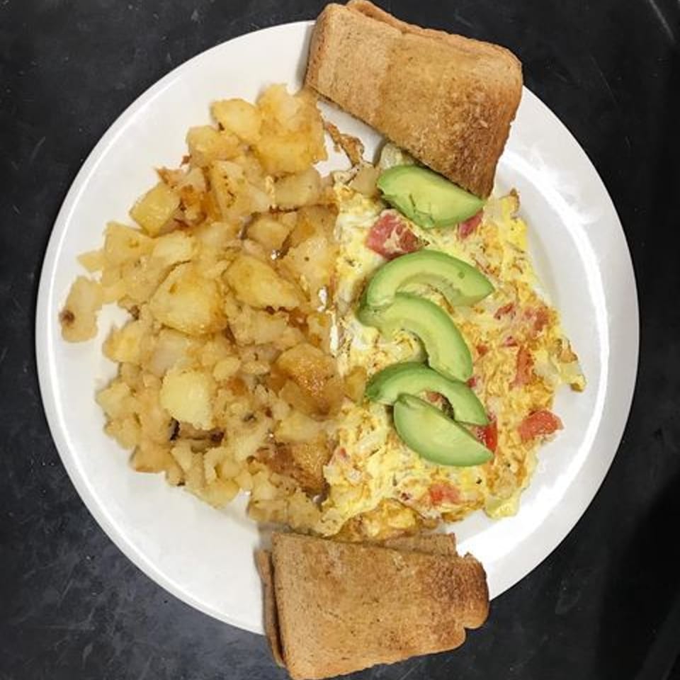 A diner breakfast plate with scrambled eggs, home fries, avocado slices, and toast.